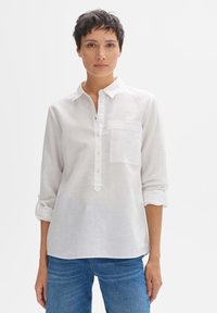 Camicia bianca in lino con bottoni, tasca sul petto e maniche arrotolate. Abbinata a jeans blu con vestibilità comoda.