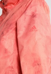 Lähikuva henkilöstä, jolla on päällään punainen Adidas-takki, jossa on hienovarainen kuvioitu tekstuuri ja Adidas-logo ylävarressa.