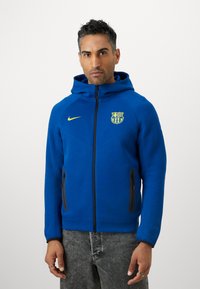 Μπλε φούτερ με φερμουάρ και κουκούλα, με λογότυπο της Μπαρτσελόνα και κίτρινο σήμα της Nike. Περιλαμβάνει μαύρες τσέπες με φερμουάρ και υφαντό ύφασμα.