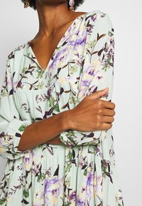 Sukienka w kwiaty z mięty zielonej tkaniny z dekoltem w szpic, ozdobiona fioletowymi i żółtymi kwiatami, z długimi rękawami i rozszerzanym dołem z marszczeniami.