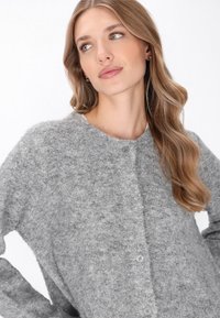 Femeie cu păr lung și ondulat blond, îmbrăcată într-un cardigan gri confortabil cu nasturi, privindu-se ușor spre stânga pe un fundal alb.