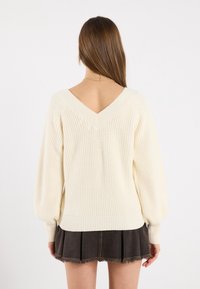 Pull en tricot crème avec un col en V et un motif côtelé texturé, associé à une jupe plissée gris foncé. Le pull a des manches longues légèrement bouffantes.