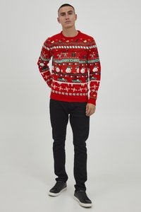 Roter gestrickter Weihnachtspullover mit Mustern von Schneemännern, Autos und Zuckerstangen. Kombiniert mit schwarzen Hosen und schwarzen Sneakern.