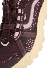 Gros plan d'une chaussure Vans marron foncé avec une semelle beige, dotée de lacets épais, de détails en daim et d'un marquage blanc audacieux sur le panneau latéral.