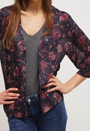 Nainen pukeutuneena tummaan kukalliseen avoimeen neuletakkiin, harmaan V-kaula-aukkoinen paita ja siniset farkut, kädet lepäävät lanteilla.