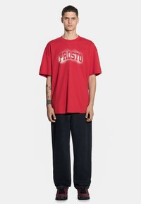 Czerwona, oversize'owa koszulka z nadrukiem "PROSTO" na białym tle na piersi, w połączeniu z luźnymi czarnymi dżinsami i czarno-czerwonymi trampkami.