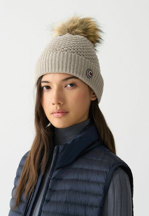 Lichtbeige gebreide muts met een ribbelrand, voorzien van een bontpompon bovenop en een klein rond logo aan de zijkant.