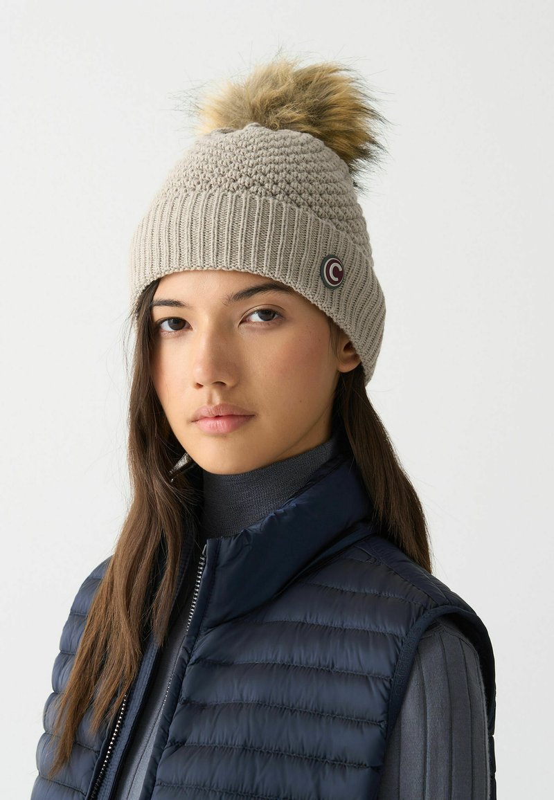 Berretto lavorato a maglia di un beige chiaro con fondo a costine, dotato di un pompon in pelliccia in cima e un piccolo logo circolare sul lato.