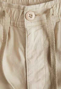 Los pantalones cortos de algodón beige cuentan con una cinturilla elástica con cordón y un único botón de color claro, mostrando una textura suave y costuras limpias.