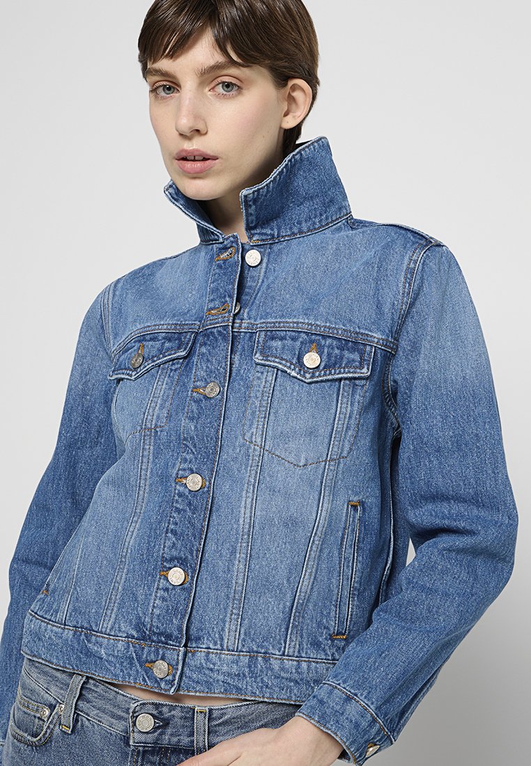 Helle blaue Denimjacke mit hohem Kragen, Fronttaschen und Metallknöpfen. Besitzt ein tailliertes Design und strukturiertes Material.