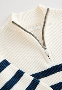 Maglione a coste color crema con collo alto, dotato di una zip metallica e strisce blu navy sulle maniche. Etichetta del marchio visibile all'interno.