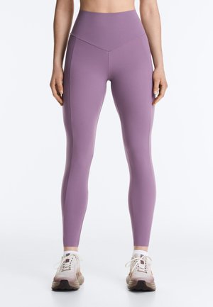 Vrouw in hooggetailleerde lavendelkleurige leggings en witte sportschoenen, staand met armen ontspannen langs het lichaam tegen een effen achtergrond.