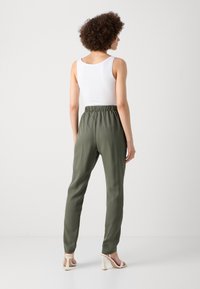 Olivgrüne Hose mit elastischem Bund, schmal zulaufenden Beinen und glatter Textur, kombiniert mit einem eng anliegenden weißen Tanktop.