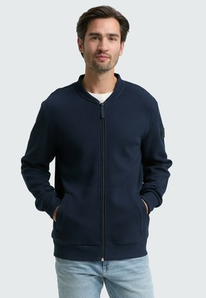 Marineblå zip-up jakke med struktureret finish, rund krave og sidelommer. Har ribbede mansjetter og nederste kant. Bæres over en lys skjorte.