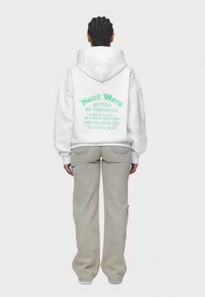 Witte oversized hoodie met groene tekst op de achterkant die zegt "Next Wave Better Be Prepared" met uitgebreide zinnen. Gdragend met lichtgrijze wijde jeans.