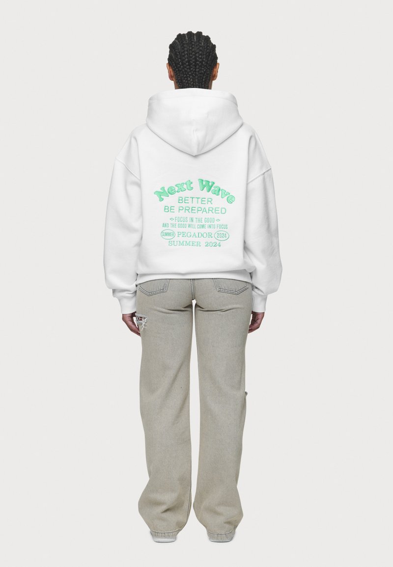 Bílá oversized mikina s kapucí, na zadní straně sytě zelený text "Next Wave Better Be Prepared" s podrobnými frázemi. V kombinaci se světle šedými širokými džíny.