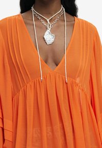 Abito di chiffon arancione con texture plissettata e profondo scollo a V, accompagnato da una collana con pendente argento su un cordino bianco.