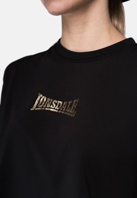 Black t-shirt featuring a metallic gold "Lonsdale" logo. The fabric is smooth with a crew neck and visible stitching details.