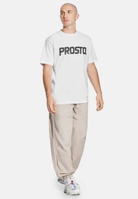 Biała, krótki rękaw t-shirt z wyraźnym czarnym napisem "PROSTO."; w połączeniu z luźnymi beżowymi spodniami cargo i białymi sneakersami. Prosty design, casualowy krój.