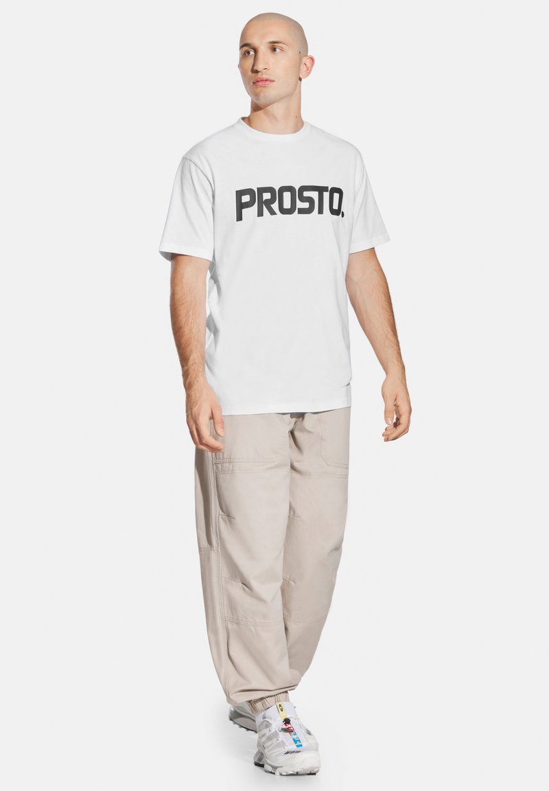 Biała, krótki rękaw t-shirt z wyraźnym czarnym napisem "PROSTO."; w połączeniu z luźnymi beżowymi spodniami cargo i białymi sneakersami. Prosty design, casualowy krój.