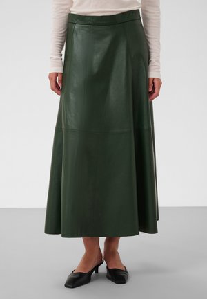 Mujer con falda midi de cuero verde oscuro, camiseta blanca de manga larga y zapatos negros de tacón bajo y punta puntiaguda, de pie sobre un suelo gris.