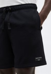 Μαύρα σορτς Björn Borg με ρυθμιζόμενο κορδόνι στη μέση και πλαϊνές τσέπες, φορεμένα από άτομο με σκουρόχρωμο δέρμα, με το χέρι στην τσέπη, σε λευκό φόντο.