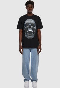 Černé bavlněné tričko s velkým, detailn�ím potiskem lebky v šedé a modré barvě. V kombinaci se světle modrými volnými džíny a bílými teniskami.