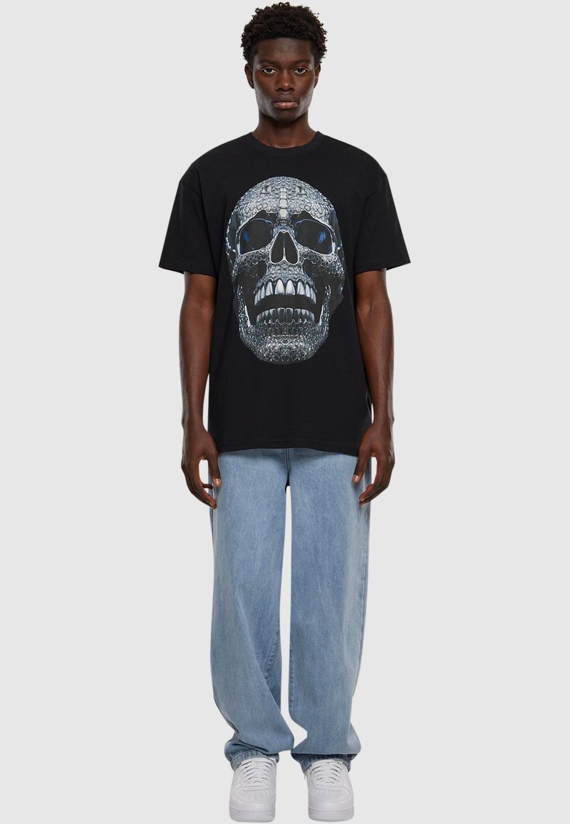 Musta puuvillapaita, jossa on suuri, yksityiskohtainen pääkallo graafinen kuva harmaana ja sinisenä. Yhdistetty vaaleansinisiin väljään farkkuihin ja valkoisiin tenniskenkiin.