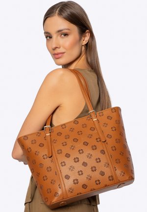 Frau mit langem braunem Haar, trägt ein ärmelloses olivgrünes Oberteil und eine große braune Ledertasche mit Muster über der Schulter.