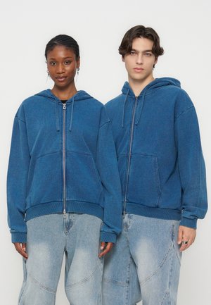 To unge voksne iført blå hættetrøjer med lynlås og lyseblå vide jeans, stående side om side mod en ensfarvet baggrund.