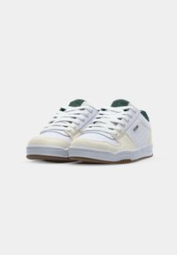 Adidași albi cu un vârf din suede off-white texturat, accente verzi pe limba pantofului și o talpă din cauciuc cu un model de aderență.