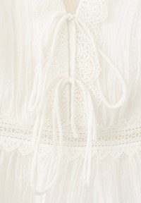 Tissu blanc avec une bordure en dentelle délicate et deux nœuds noués avec des cordons fins sur un vêtement texturé.