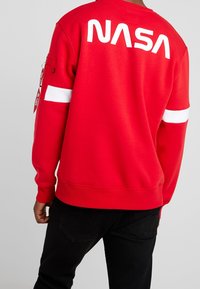 Felpa rossa con logo "NASA" bianco sulla schiena; presenta una striscia bianca sulle maniche e una tasca laterale con zip.