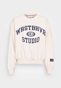 Sudadera beige con texto en negrita azul marino "WRSTBHVR STUDIO" y "03D" en un óvalo en la parte frontal, puños y dobladillo acanalados, y un pequeño parche en la manga izquierda.