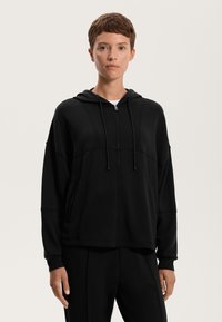 Fekete zipzáras kapucnis pulóver, húzózsinórral, amely lazán illeszkedik, oldalsó zsebekkel és diszkrét varrási részletekkel rendelkezik. Puha anyagú textúra.