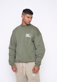 Olijfgroen sweatshirt met een ronde hals en een klein wit logo op de borst. Heeft loszittende mouwen en een zachte textuur.