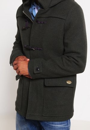 Homme portant un manteau à brandebourgs vert foncé par-dessus une chemise bleue et un jean, les mains jointes devant, sur un fond clair uni.