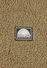 Brun fleece tekstil med en tekstureret overflade og en sort etiket, der har en hvid regnbue og teksten "SOUTHPOLE ORIGINALS."