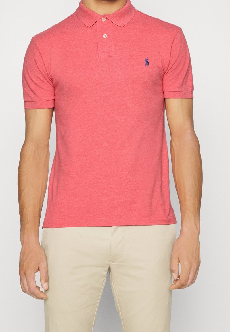 Homme portant un polo corail à manches courtes avec deux boutons et un pantalon beige, debout devant un fond uni.