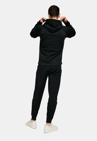 Haut à capuche noir et pantalon de survêtement assorti en tissu doux. Comprend une capuche, des poignets côtelés et un détail de logo près de la cheville. Porté avec des baskets blanches.