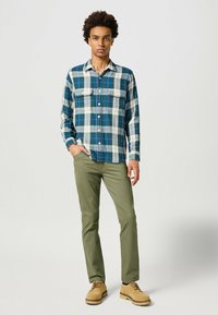 Camisa de franela a cuadros en azul, verde y blanco, con dos bolsillos en el pecho, cuello abotonado, combinada con pantalones verde oliva y zapatos marrón claro.
