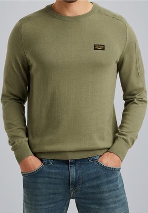 Homme portant un pull à manches longues vert olive avec un écusson logo PME Legend et un jean bleu, mains dans les poches, sur un fond gris uni.