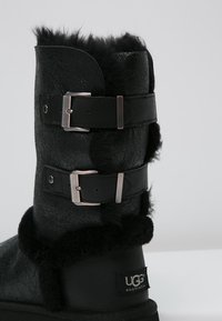 Botte en cuir noir avec deux sangles à boucles, présentant une bordure en fourrure noire douce en haut et un logo UGG embossé sur le côté.