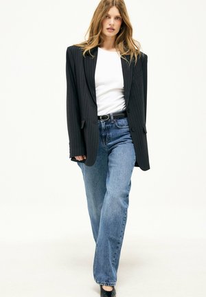 Woman with long hair wearing a black pinstripe blazer over a white shirt, blue jeans, and black belt, walking forward on a white background.