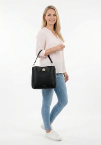 Šypsanti moteris šviesios palaidinės ir džinsų dėvėdama didelę juodą rankinę ant peties, stovinti priešais baltą foną.