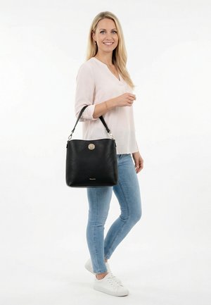 Lachende vrouw in een lichte blouse en spijkerbroek, met een grote zwarte handtas op haar schouder, staand tegen een witte achtergrond.