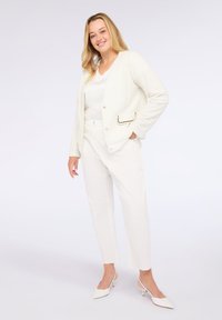 Mujer sonriente de pie, vestida con un conjunto completamente blanco que incluye una chaqueta texturizada, blusa blanca, pantalones sastre y tacones afilados, frente a un fondo liso.