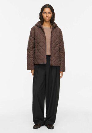 Donna che indossa una giacca trapuntata marrone con cappuccio sopra un maglione beige, pantaloni neri a gamba larga e scarpe scure, in piedi su uno sfondo chiaro.