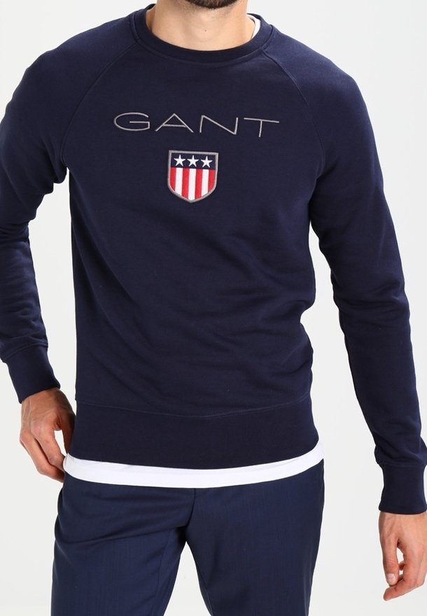 Homme portant un sweat-shirt GANT bleu marine avec un logo en forme de bouclier affichant trois étoiles et des rayures rouges et blanches, assorti à un pantalon bleu marine.