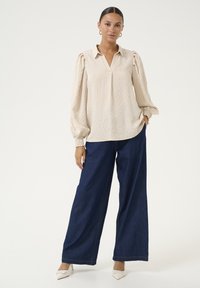 Vrouw in een crèmewitte blouse met lange mouwen en pofmouwen, donkere wijde jeans, witte hakken en gouden oorringen, die met één hand in haar zak staat.